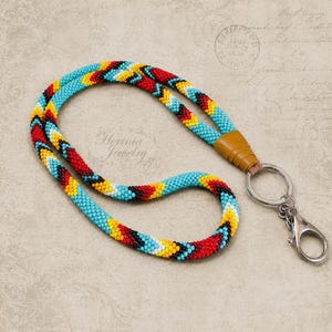 May include: A beaded lanyard featuring a repeating chevron pattern in turquoise, red, yellow, and black glass beads. The cord is secured with a tan leather connector and includes a silver-toned metal split ring and a lobster claw clasp attachment.
