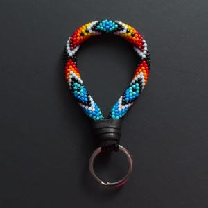 May include: A beaded keychain with a colorful pattern of red, orange, yellow, blue, and black beads. The keychain has a black leather loop and a silver key ring. The text at the bottom says, "Please, respect my artistry and do not copy or imitate!"