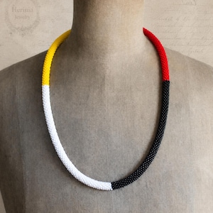 May include: A long, tubular beaded necklace featuring a color-blocked design. The necklace is composed of small, uniform beads in four distinct sections of yellow, white, red, and black, arranged in a continuous, smooth, and flexible circular strand.