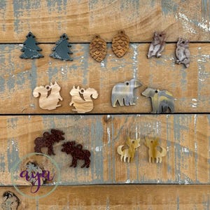 May include: A collection of small, flat, nature-themed charms in various shapes including pine trees, pinecones, owls, squirrels, bears, moose, and foxes. The charms feature diverse textures and colors, including dark green, brown, and marbled patterns.