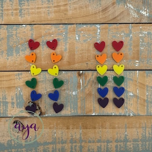May include: A rainbow of wooden heart-shaped charms, arranged in two columns, on a wooden background. The hearts are red, orange, yellow, green, blue, and purple.