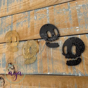 May include: Four skull-shaped pendants arranged in a row. Two are translucent gold with embedded glitter, and two are solid black with a detailed lace-like pattern cut into the surface. Each skull features two eye sockets and a separate jaw piece.