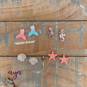 May include: A set of colorful resin mermaid tail, seahorse, seashell, and starfish charms. The mermaid tail charms are iridescent and have a pink and blue color shift. The seahorse charms are pink with glitter. The seashells are white with a pearlescent finish. The starfish charms are coral pink. The charms are arranged on a wooden surface. **IRIDESCENT COLOR SHIFT** is written in white text on a black background.