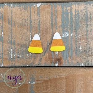 May include: A pair of candy corn earrings. The earrings are made of felt and have a white, orange, and yellow design.
