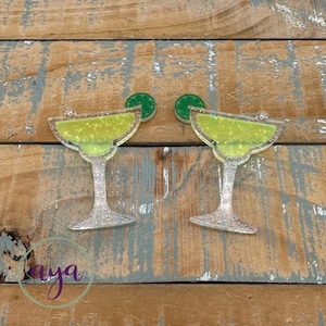 May include: Two clear plastic margarita glasses with green glitter and a lime wedge on top. The glasses are on a wooden background.