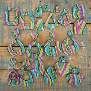 May include: A collection of colorful wooden earrings in various shapes, including squares, triangles, and teardrops. The earrings feature a vibrant, abstract design with pink, yellow, and teal stripes. The earrings are arranged on a weathered wooden surface.