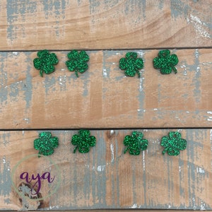 May include: Eight green glitter four-leaf clover charms on a wood background.
