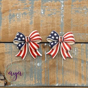 May include: Two patriotic bows with a red, white, and blue color scheme. The bows feature a design reminiscent of the American flag, with stars and stripes. The bows are likely for crafting or decorative purposes.