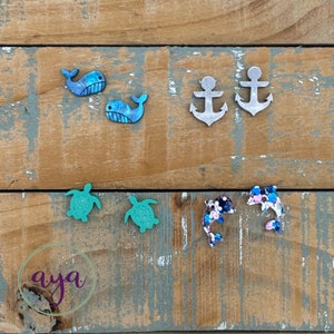 May include: A set of 10 colorful resin earrings featuring various sea creatures, including whales, anchors, turtles, and dolphins. The earrings are displayed on a wooden background.