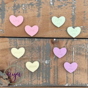 May include: Eight heart-shaped candy conversation hearts with various messages, including "Love", "Cutie Pie", "Dream", and "Be Mine". The hearts are in pink, light green, yellow, and light purple.