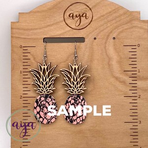May include: A pair of pineapple-shaped earrings with a pink and black patterned design. The earrings are approximately 3 inches long and are displayed on a wooden board with a ruler.