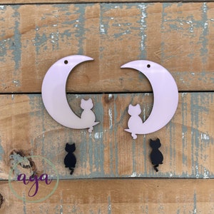 May include: Two pink crescent moon shaped earrings with black cat silhouettes. Each earring has a black cat silhouette sitting on the moon. There are two additional black cat silhouettes.