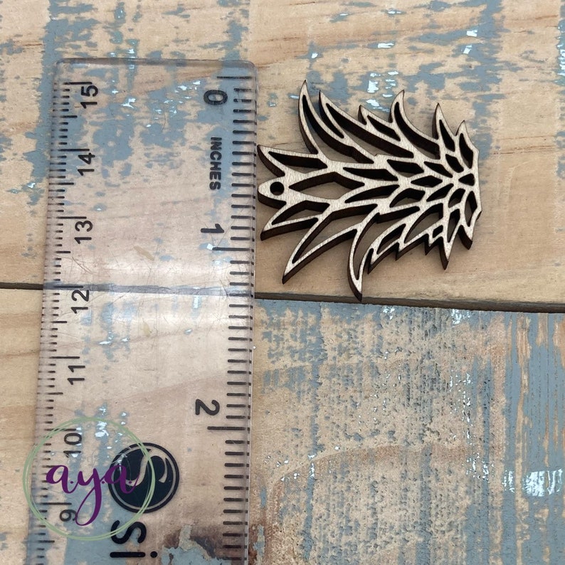 May include: A wooden cutout of a stylized pineapple with a hole at the top for hanging. The cutout is approximately 2 inches tall and is shown next to a ruler.