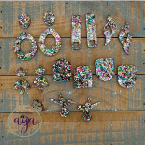 May include: Assortment of colorful confetti resin earrings in various shapes, including ovals, rectangles, and abstract shapes. The earrings are displayed on a wooden surface.