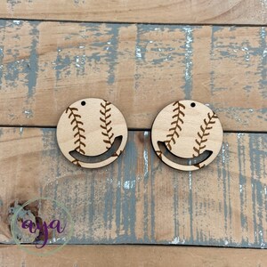 May include: Two wooden earrings shaped like baseballs with brown stitching. The earrings are unfinished and ready to be painted or stained.