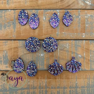 May include: A collection of wooden earrings in various shapes, including teardrops, ovals, and circles. The earrings feature a blue, purple, and white leaf pattern. The earrings are displayed on a weathered wooden surface.