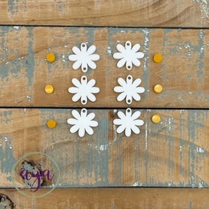 May include: Six white daisy-shaped charms with a hole in the centre, and six small yellow circles on a wooden background.