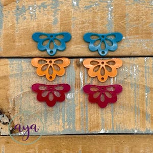 May include: Six decorative floral-shaped embellishments in blue, orange, and red-pink hues. They are arranged on a weathered wooden surface. The word "aya" is in the lower left corner.