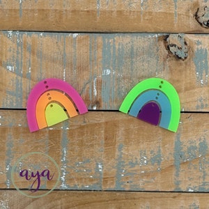 May include: Two rainbow-shaped acrylic charms, one in shades of pink, orange, and yellow, and the other in shades of green, blue, and purple. Both charms have three layers and are decorated with small holes.