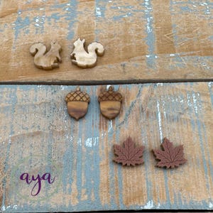 May include: Wooden fall-themed cutouts including two squirrels, two acorns, and two maple leaves. The cutouts are in shades of brown and are arranged on a rustic, weathered wooden surface. The word "aya" is written in cursive.