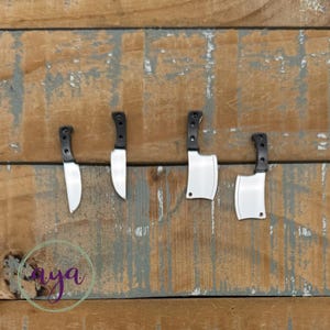 May include: Four miniature kitchen knives with black handles and silver blades are displayed against a weathered wooden background. The knives include two small paring knives and two cleavers. The image also includes the word "aya" in a circular design.