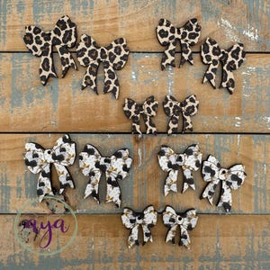 May include: Wooden bows in various sizes and patterns, including leopard print and a black and white floral design. The bows are arranged on a rustic, weathered wooden surface, showcasing their decorative appeal.