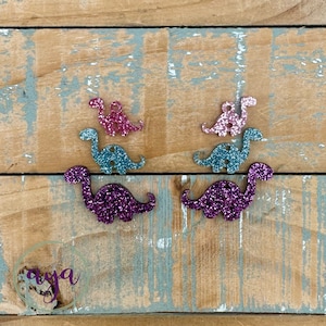 May include: A set of six glitter dinosaur shapes in pink, teal and purple. The dinosaurs are arranged in pairs, with a smaller dinosaur on top of a larger dinosaur.