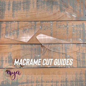 May include: Two clear plastic macrame cut guides on a wooden surface. The guides are shaped like triangles and curved lines. The text "MACRAME CUT GUIDES" is printed on the wood surface.