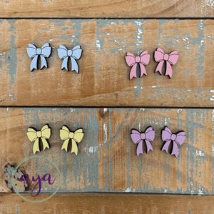 May include: Six wooden bow tie shapes in various pastel colors. The bows are arranged in two rows of three. The colors are light blue, pink, yellow, and lavender.