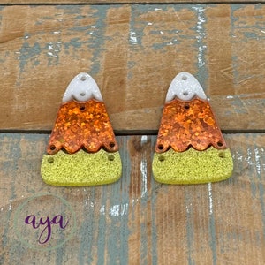 May include: A pair of candy corn shaped earrings with glitter. The earrings are white, orange, and yellow.