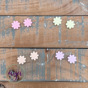 May include: Assortment of pastel colored flower shaped buttons in pink, light purple, and yellow. The buttons are arranged in two rows on a wooden surface.