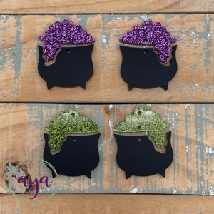 May include: Four black cauldron ornaments with glitter accents. Two cauldrons have purple glitter and two have green glitter.