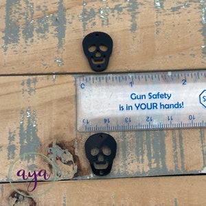 Puede incluir: Dos abalorios de acr&iacute;lico en forma de calavera negros con detalles recortados. Los abalorios miden aproximadamente 2,5 cm y se muestran junto a una regla.