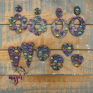 May include: A collection of colorful skull-themed earrings and pendants. The earrings include round studs, flower shapes, and oval hoops. The pendants are heart-shaped, skull-shaped, and guitar pick-shaped. Each piece features a repeating skull pattern with vibrant colors.