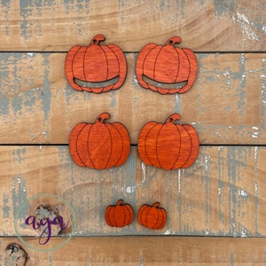 May include: Five wooden pumpkin cutouts in various sizes. The pumpkins are painted a bright orange colour and have a wood grain texture. The pumpkins are arranged in a staggered pattern on a wooden surface.