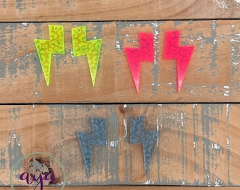 May include: Three pairs of lightning bolt shaped earrings. The earrings are made of clear acrylic with a leopard print pattern. The earrings are in three colors: yellow, red, and blue.