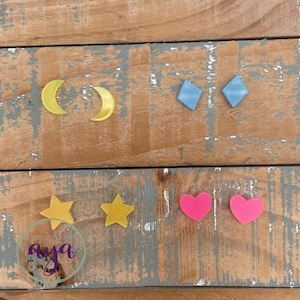 May include: A set of six colorful earrings featuring a crescent moon, a diamond, a star, and a heart. The earrings are made of acrylic and are available in yellow, blue, and pink.