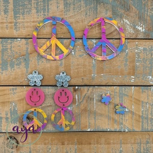 May include: Assortment of colorful peace sign and smiley face earrings. The earrings are made of resin and have a tie-dye effect. There are two pairs of peace sign earrings, two pairs of smiley face earrings, and two pairs of flower earrings.