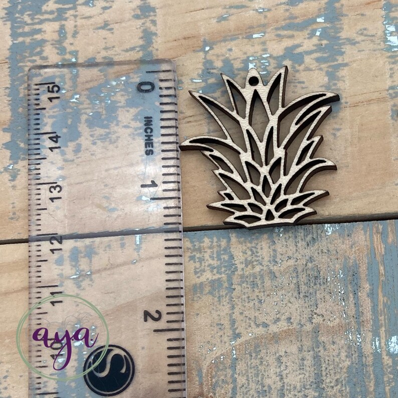 May include: A wooden cutout of a pineapple with a hole at the top for hanging. The cutout is about 1 inch tall and is shown next to a ruler.
