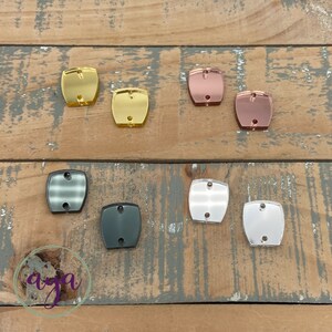 May include: Assortment of eight square-shaped acrylic mirror tiles in various colors, including gold, rose gold, silver, black, and clear. The tiles have two holes in the top for hanging or attaching.