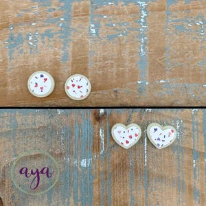 May include: Four wooden ornaments with a white background and pink, red, and purple confetti. Two ornaments are round and two are heart shaped.