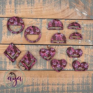 May include: Assortment of wooden earring blanks with a floral pattern in shades of pink and purple. The blanks are in various shapes, including circles, hearts, squares, and half-circles.
