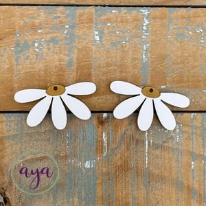 May include: Two white wooden daisy earrings with yellow centers. The earrings are on a wooden background.