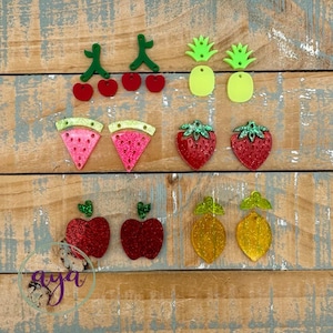 May include: A collection of colorful fruit-shaped acrylic charms, including cherries, pineapples, watermelon slices, strawberries, apples, and lemons. The charms are glittery and have green accents. The background is a weathered wooden surface.