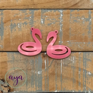 May include: Two pink flamingo-shaped cutouts with a small hole at the top for hanging. The cutouts are displayed on a weathered wooden surface. The word "aya" is written in cursive in a circle in the lower left corner.