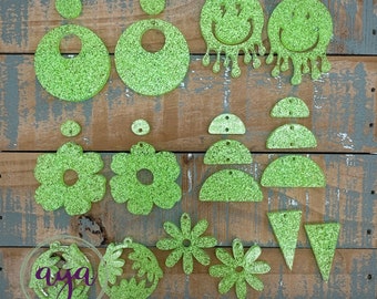 May include: A collection of lime green glitter acrylic earring components in various shapes, including circles, flowers, triangles, and smiley faces. Some pieces have holes for attaching to earring hooks. The word "aya" is visible in a clear circle.