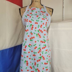 May include: A light blue and white gingham apron with a pattern of red cherries and green leaves. The apron is displayed on a mannequin and has a light blue neck strap and ties.