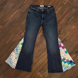 May include: Dark wash denim jeans with patchwork bell bottoms. The patchwork features a variety of colorful fabric squares in shades of blue, green, pink, and purple. The jeans have a classic five-pocket design.