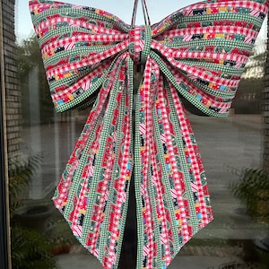 May include: A large, decorative bow with a festive design. The bow features a red and white checkered pattern, accented with green trim and a repeating pattern of Christmas-themed elements. It is hanging on a window.
