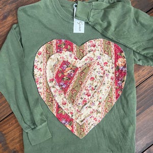 May include: An olive green long-sleeve shirt featuring a large, layered heart appliqué on the front. The heart is constructed from three stacked fabric layers with a floral and striped pattern in shades of red, cream, and green. A white tag reads Grace.